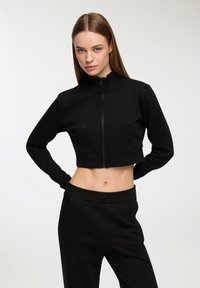 Felpa nera corta con colletto alto e chiusura a zip, dotata di tasca frontale e texture morbida. Abbinata a pantaloni della tuta neri coordinati.
