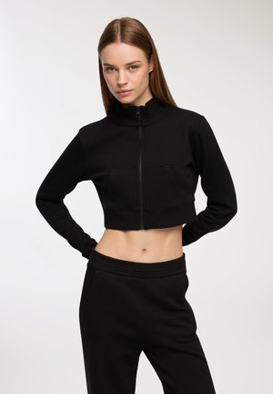 Czarna, krótka bluza z wysokim kołnierzem i zapięciem na zamek, wyposażona w przednią kieszeń i miękką teksturę. W połączeniu z pasującymi czarnymi spodniami dresowymi.