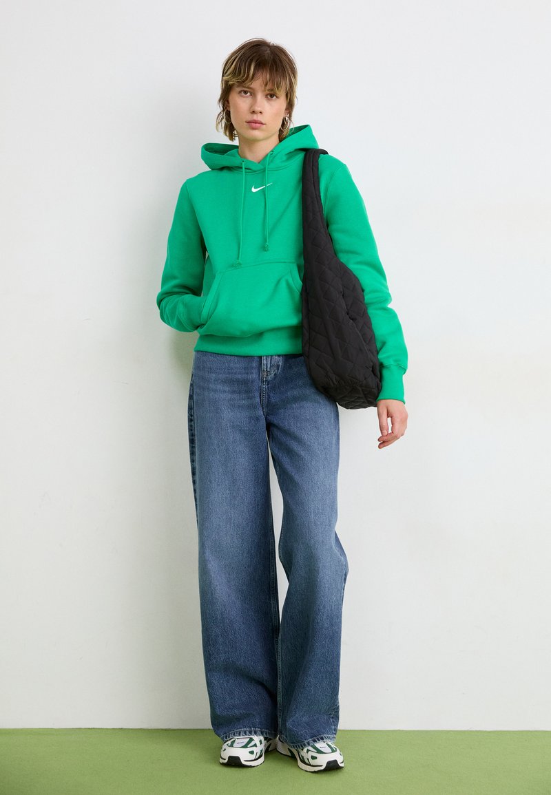 Sweat à capuche vert avec une poche avant, associé à un jean bleu à jambe large. Sac à bandoulière noir matelassé. Baskets blanches avec des accents verts.