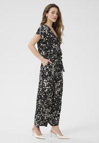 Svart blommig jumpsuit med korta ärmar, knytmidja och fickor. Har ett mönster med vita blommor. Modellen bär ljusbeige slingback-hälskor.