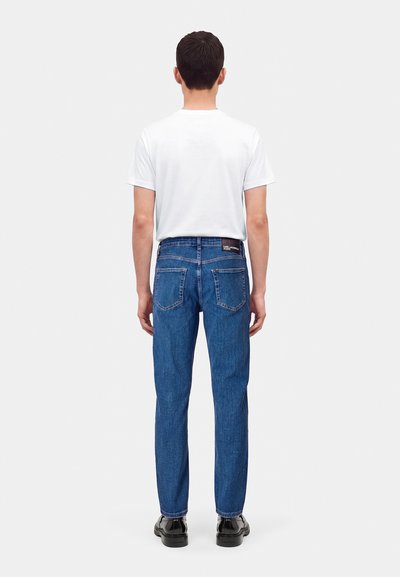 Homme debout de dos portant un t-shirt blanc uni, un jean bleu avec une étiquette à la taille, et des chaussures noires sur fond blanc.
