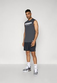 Camiseta cinzenta sem mangas com o texto "RAIDERS Football", combinada com calções escuros. Usada com meias brancas da Nike e ténis preto e branco.