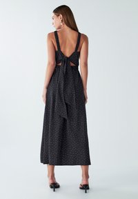 Žena stojí, oblečená v čiernych bezrukávových midi šatách s bielymi bodkami, s otvoreným chrbtom viazaným mašľou, a v čiernych sandáloch na opätku.