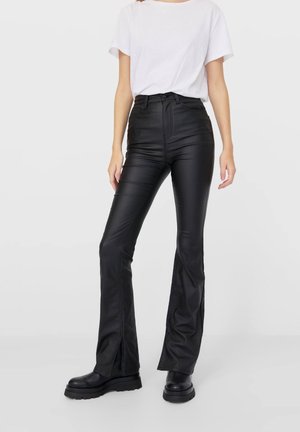 Person trägt schwarze High-Waist-Lederhose mit weitem Bein und vorderen Schlitzen, kombiniert mit einem schlichten weißen Kurzarmhemd und schwarzen klobigen Stiefeln.