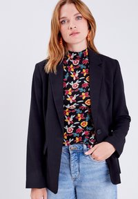 Blazer negro entallado con dos botones, llevado sobre una blusa de estampado floral que presenta flores rojas, amarillas y azules sobre un fondo oscuro.