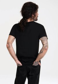 Camiseta negra de algodón con mangas cortas, escote redondeado y un corte ajustado, combinada con jeans negros. Tatuajes visibles en los brazos.