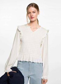 Biela blúzka s dlhými rukávmi, riasenými detailmi, smotaným živôtikom a rozšíreným spodným okrajom. V kombinácii so svetlomodrými nohavicami a držiacim tmavomodrým sakom.