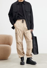 Svart zip-up jacka över en svart skjorta, beige cargo-byxor med sidofickor, svarta snörstövlar, håller en svart tote-väska. En enfärgad bakgrund.