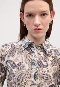 Camisa colarinho em creme claro com padrão paisley azul e bege, apresentando a bainha arredondada e fecho com botão, feita de tecido suave.