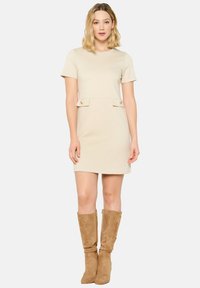 Beige Kurzarmkleid mit eng anliegendem Oberteil, zwei Fronttaschen und goldfarbenen Knöpfen. Kombiniert mit knielangen tanfarbigen Wildlederstiefeln.