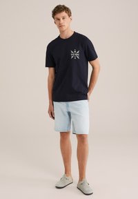 Navy kokvilnas T-krekls ar priekšējo grafiku, uz kura rakstīts "BLUE RIDGE", savienots ar gaišiem džinsa šortiem un pelēkiem slip-on apaviem.