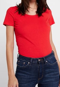 Top corto rojo de manga corta, hecho de una tela suave, con un escote redondo, combinado con unos pantalones vaqueros de denim azul oscuro que presentan herrajes en tono cobre y bolsillos.