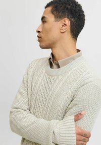 Pull en tricot beige clair avec un motif texturé, des poignets côtelés et un col rond, superposé à une chemise à col beige.