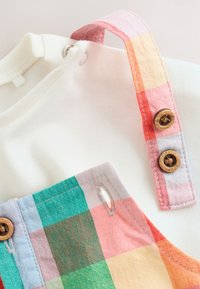 Vêtement blanc pour bébé avec une sangle à carreaux arc-en-ciel pastel, maintenue par deux boutons marron sur un fond clair.