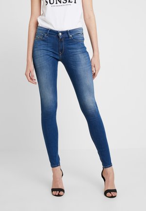 Vrouw draagt blauwe skinny jeans gecombineerd met zwarte open-toe hakken en een wit topje met de tekst „SUNSET“, staand tegen een effen achtergrond.