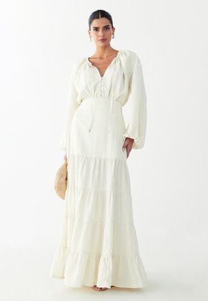 Vrouw met donker haar in een lange crèmekleurige jurk met opgeblazen mouwen, ze houdt een rond gevlochten tas vast tegen een effen witte achtergrond.