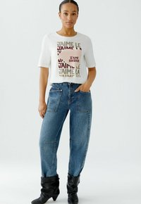 Weißes T-Shirt mit kurzen Ärmeln und mehrfarbigem Text, kombiniert mit lockerer blauer Jeans und schwarzen, lässigen Stiefeletten.