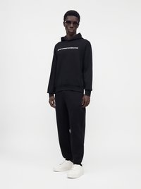 Άνδρας που φοράει μαύρο φούτερ Emporio Armani και μαύρο παντελόνι με λευκά αθλητικά παπούτσια, στέκεται μπροστά σε απλό λευκό φόντο.