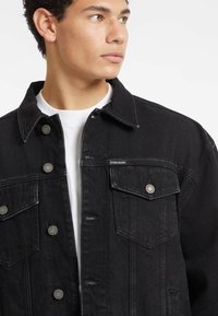 Giacca di jeans nera con taglio classico, frontale con bottoni, due tasche sul petto e dettagli in hardware argentato. Indossata sopra una camicia bianca.