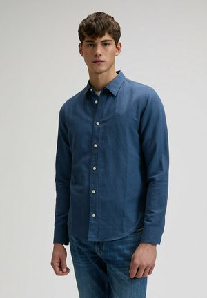 Młody mężczyzna ubrany w niebieską koszulę z długim rękawem na guziki i niebieskie dżinsy, stojący na jasnym, jednolitym tle.