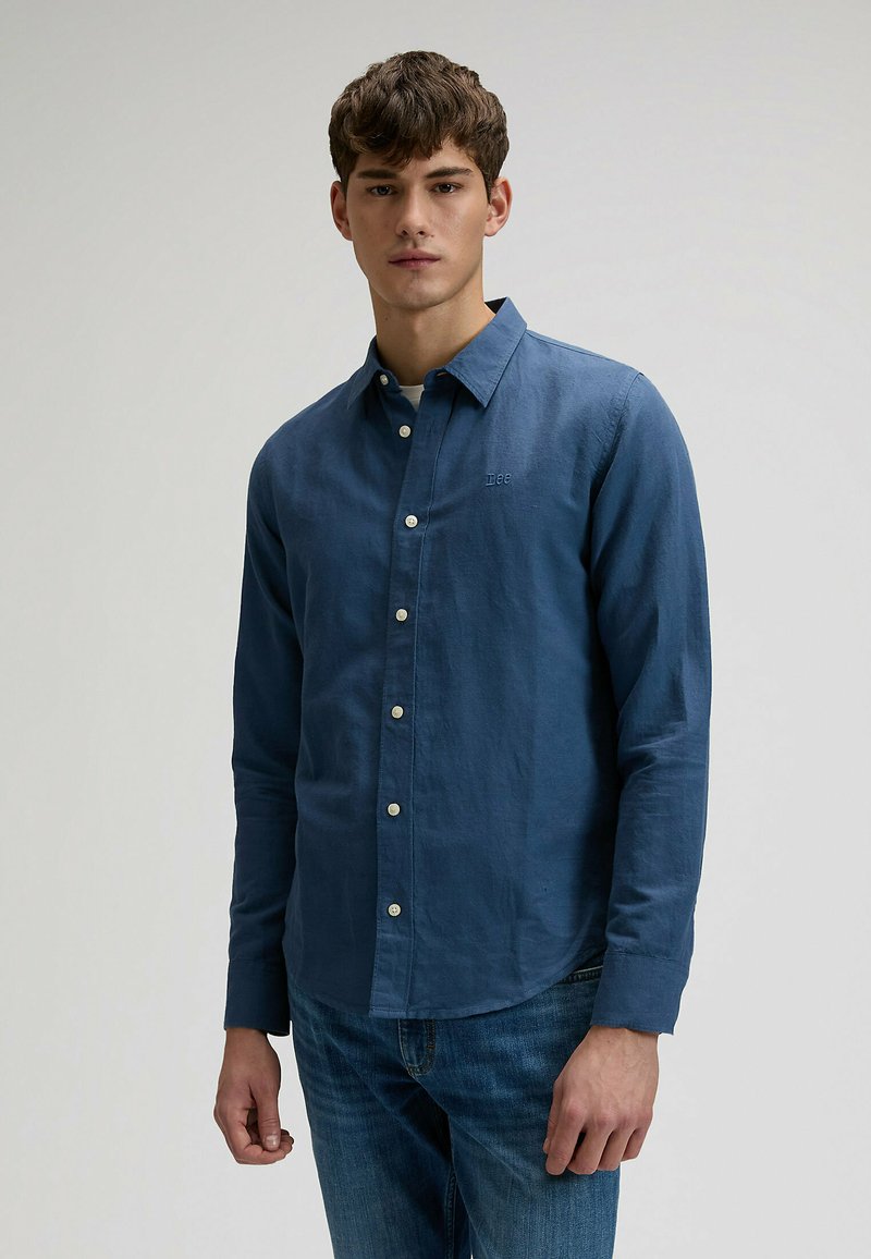 Junger Mann trägt ein blaues langärmliges Hemd mit Knöpfen und eine blaue Jeans, steht vor einem schlichten, hellen Hintergrund.