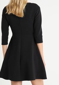 Svart klänning med en figurnära bodice, trekvartärmar och en utställd kjol. Har ett texturerat tyg med vertikala ränder och en bakre söm.