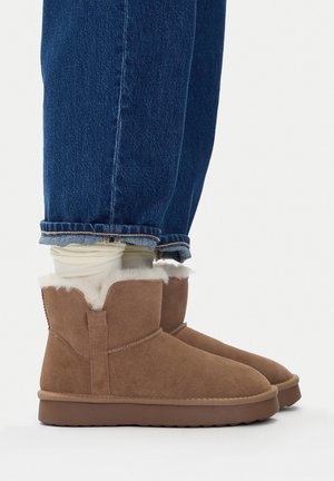 Brązowe zamszowe botki za kostkę z miękką białą podszewką z polaru, niską brązową podeszwą i przeszyciami, noszone z podwiniętymi niebieskimi dżinsami.