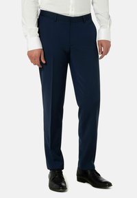 Man in donkerblauwe broek, witte lange mouwen shirt, en zwarte nette schoenen staat tegen een effen achtergrond.