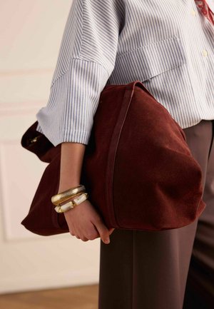 Personne portant une chemise rayée bleue et un pantalon marron tenant un grand sac en suède rouge foncé, avec des bracelets dorés et blancs au poignet.