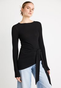 Bluză neagră cu mâneci lungi și decolteu rotund, având un detaliu cu panglică laterală. Țesătură netedă și elastică, combinată cu blugi largi, de culoare albastru deschis.