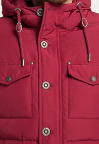 Bordeaux pufferjakke med høj krave, hætte med snore og sølvfarvede trykknapper. To klaplommer på brystet. Glat tekstur.