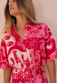 Robe chemise rose avec un motif cachemire comprenant des créatures marines et des motifs floraux. Manches courtes et ceinture à nouer à la taille.