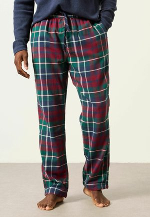 Person trägt grüne, rote und dunkelblaue karierte Pyjamahose mit den Händen in den Taschen und steht barfuß auf einem hellen Boden.