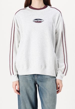 Sudadera gris claro con texto "San Diego U.S.A.", franjas burdeos en las mangas, combinada con jeans azules por una persona de pie sobre fondo blanco.