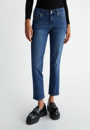 Persoon draagt blauw denim spijkerbroek met mid-rise en zwarte plateau loafers, staand tegen een effen witte achtergrond.