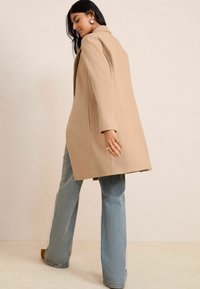Beige, op maat gemaakte overjas met gestructureerde schouders en brede mouwen, gedragen over lichtblauwe flared jeans. Soepele textuur, minimale details.
