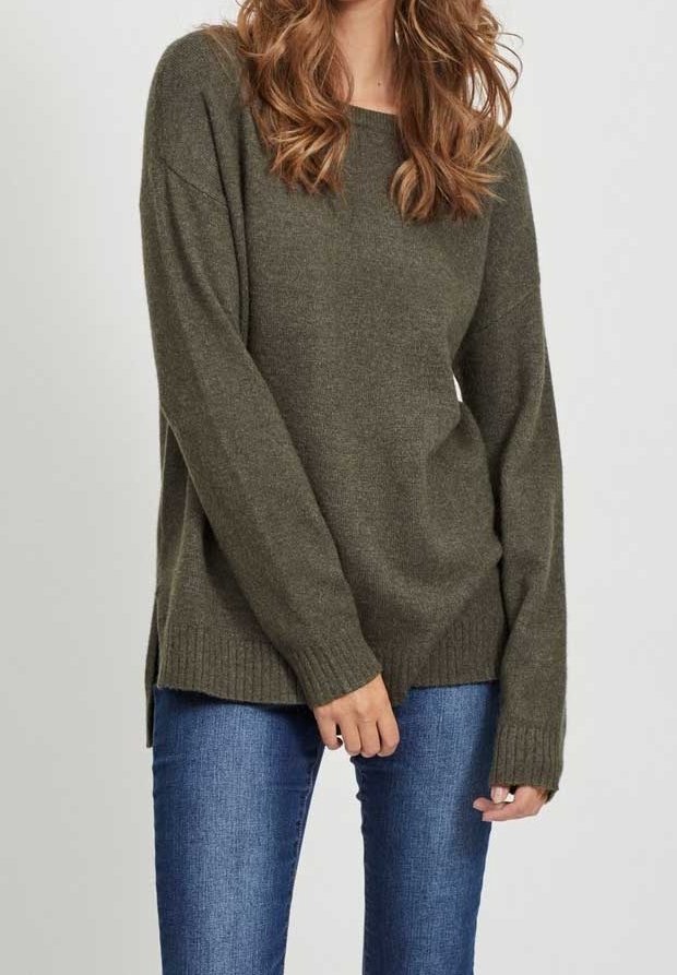 Femme portant un pull ample à manches longues vert olive et un jean bleu, debout devant un fond gris uni.