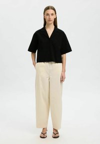Schwarzes kurzärmliges Hemd mit Knöpfen und einer einzelnen Tasche, kombiniert mit hellbeigen weit geschnittenen Hosen und flachen schwarzen Sandalen. Einfaches und modernes Design.