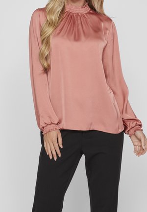 Satynowa bluzka w kolorze pudrowego różu z wysokim, falbaniastym kołnierzem i długimi rękawami zakończonymi marszczonymi mankietami. Połączona z czarnymi spodniami.