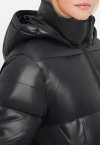 Casaco acolchoado preto com uma textura brilhante e suave, apresentando um fecho de correr e capuz. O material parece leve e isolante.