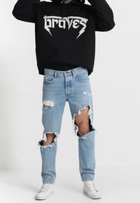 Svart huvtröja med "braves"-logga, kombinerad med ljusblå slitna jeans med stora utskärningar vid knäna. Vita sneakers fulländar outfiten.