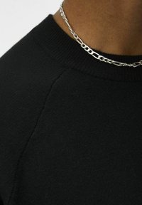 Zwarte trui met een ronde halslijn, voorzien van een gladde textuur en subtiele geribbelde details bij de kraag; gecombineerd met een zilveren ketting.