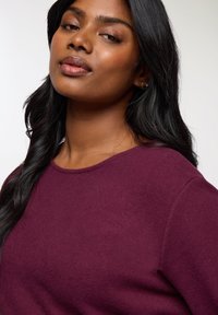 Felpa bordeaux con scollatura tonda e texture morbida. Il modello ha capelli lunghi e scuri e indossa piccole orecchini a cerchio in argento.