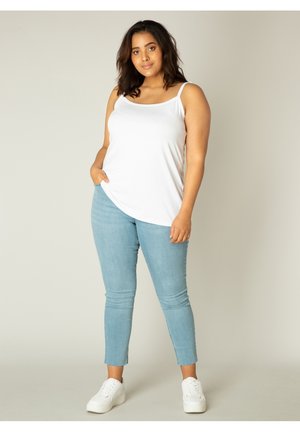 Witte mouwloze top met een ronde zoom, gecombineerd met lichtblauwe skinny jeans met een cropped lengte. Model draagt witte sneakers.