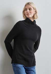 Pull à col roulé noir avec un col côtelé, fentes sur les côtés et détail de bouton. Fabriqué à partir d'un matériau doux, associé à un jean en denim.