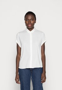 Blusa bianca a maniche corte con frontale abbottonato e colletto, caratterizzata da una texture liscia e una vestibilità morbida. Abbinata a jeans in denim scuro.