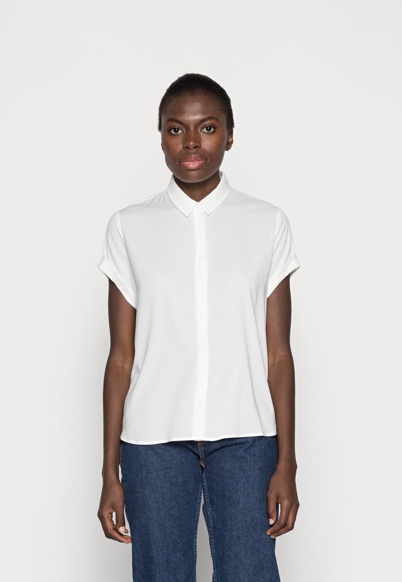 Blusa bianca a maniche corte con frontale abbottonato e colletto, caratterizzata da una texture liscia e una vestibilità morbida. Abbinata a jeans in denim scuro.