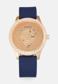 Reloj dorado con una esfera naranja brillante que presenta un patrón de triángulos, rodeada de cristales, y una correa de silicona navy.