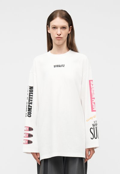 Γυναίκα που φοράει λευκό πουκάμισο μακρυμάνικο με μαύρο και ροζ κείμενο, συμπεριλαμβανομένων των λέξεων "STIEGLITZ" στο στήθος και "COMPETITION" στα μανίκια.