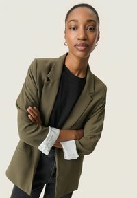 Sacou verde măsliniu cu o croială lejeră și rever ciupit, purtat peste un tricou negru. Mansetele au un accent cu dungi albe.